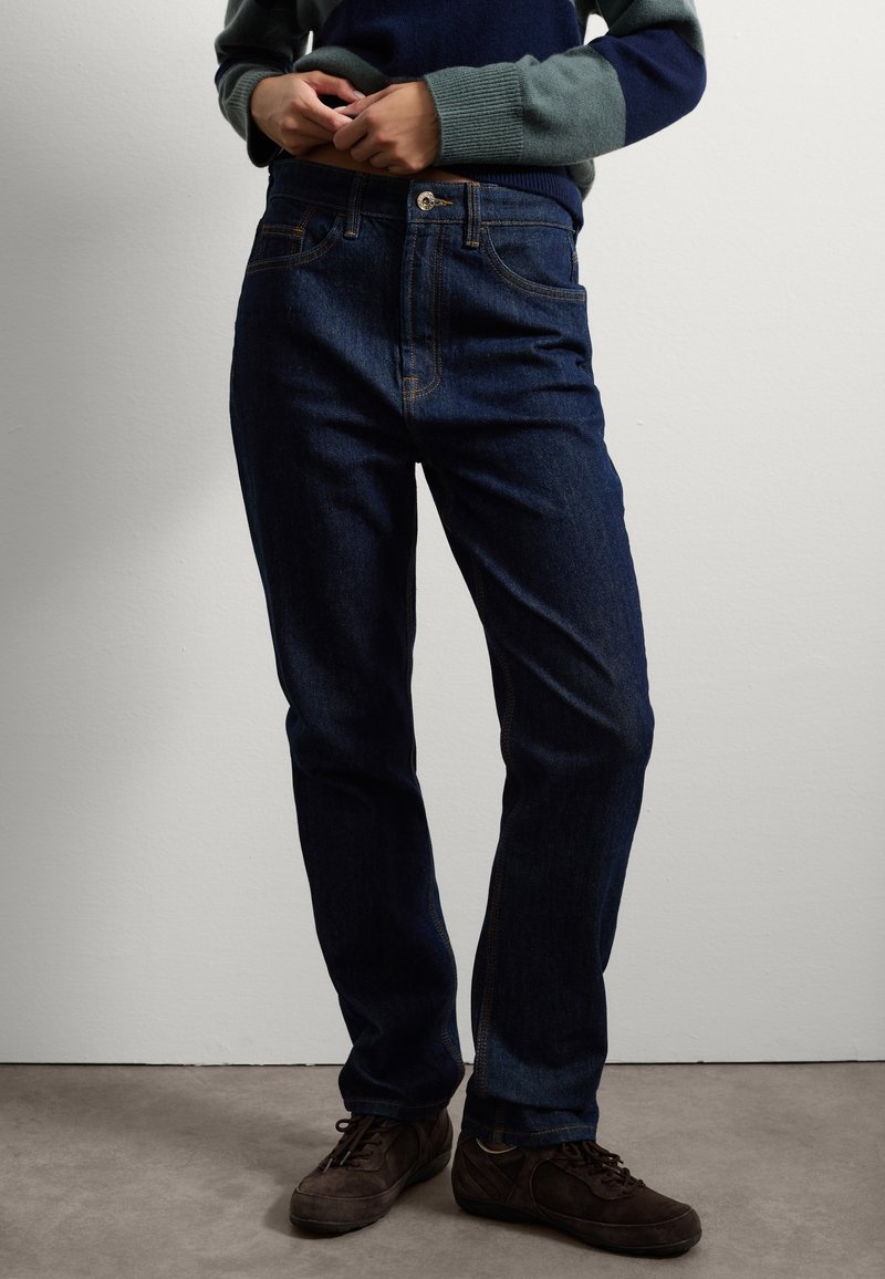 Jeans de color azul oscuro con un diseño de pierna recta, que cuentan con un cierre frontal de botón, detalles de costura clara y bolsillos. Llevados con zapatos marrones.