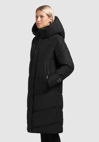 Abrigo de plumas negro con un diseño largo, capucha oversize, bolsillos con cremallera y secciones acolchadas. Fabricado con un material suave y resistente al agua.