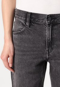 Jean en denim noir avec une finition texturée, design à cinq poches et détails de couture visibles. Fermeture à bouton et zip à l'avant.