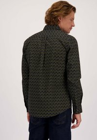 Man met golvend haar in een zwart shirt met lange mouwen en een groen abstract patroon, gecombineerd met donkerblauwe jeans, zijdelings gekeerd.