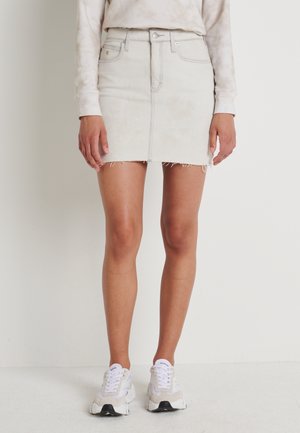 Jupe en jean mini en délavé clair avec un ourlet effiloché. Comprend cinq poches, des passants de ceinture et une fermeture à bouton à l'avant. Portée avec des baskets blanches.