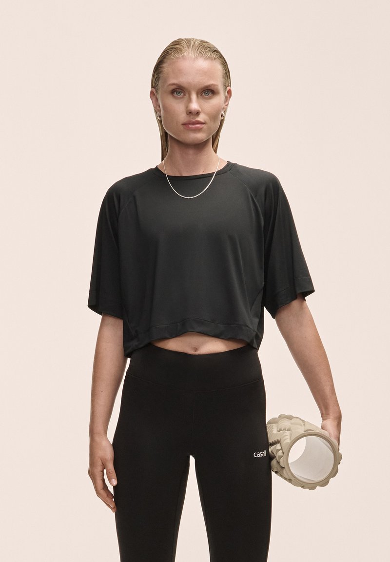 Black cropped athletic shirt with short sleeves, paired with black leggings. Model holds a textured foam roller in a light color.