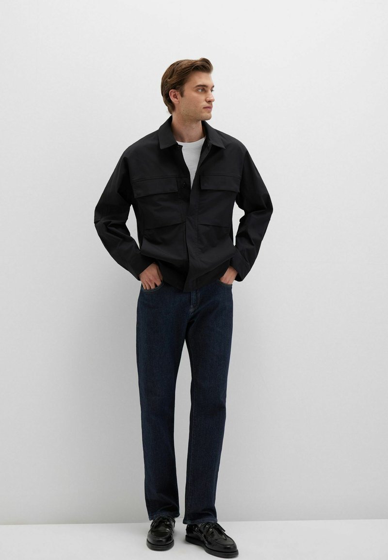 Schwarze, langärmelige Jacke mit zwei Brusttaschen, getragen über einem weißen Hemd. Kombiniert mit dunkelblauen Jeans und schwarzen Schuhen. Glatte Textur.