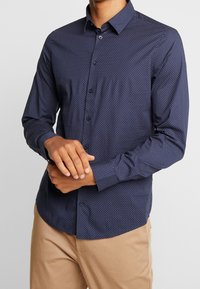 Marinblå långärmad skjorta med ett subtilt mönster av vita prickar, knappad krage och skräddarsydd passform. Avslutad med en enda bröstknapp.