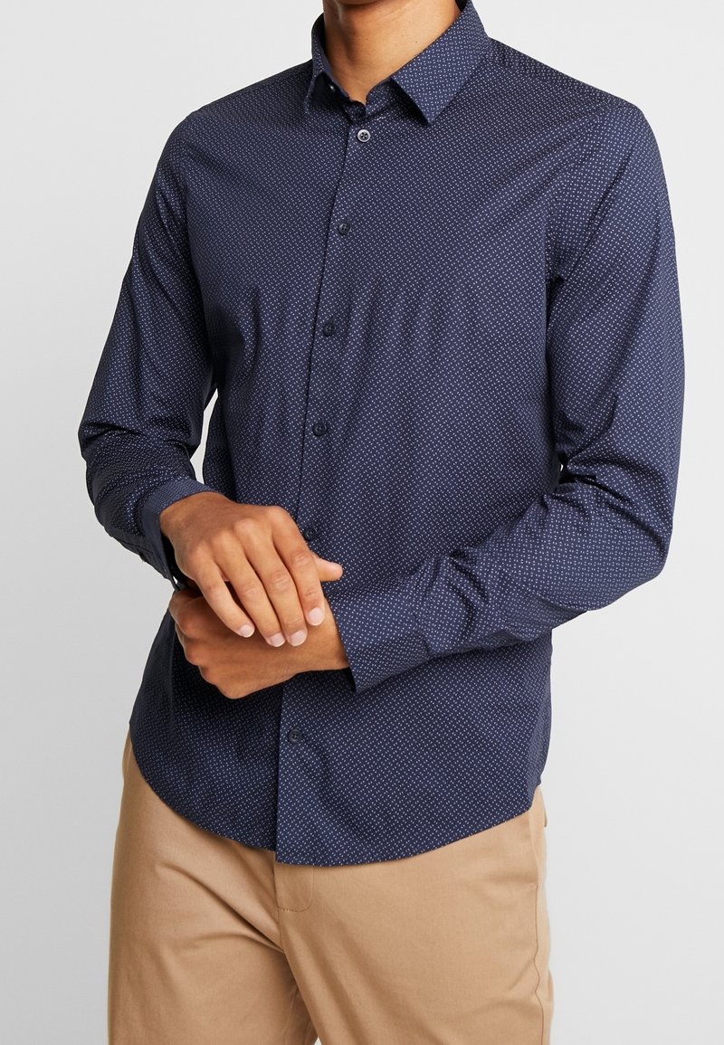 Marinblå långärmad skjorta med ett subtilt mönster av vita prickar, knappad krage och skräddarsydd passform. Avslutad med en enda bröstknapp.