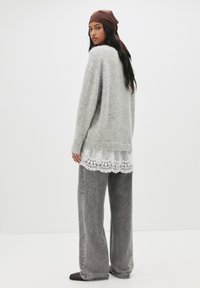 Przesadnie duży szary sweter o miękkiej fakturze, założony na biały top z koronkowym wykończeniem, zestawiony z luźnymi szarymi dżinsami i ciemnymi butami wsuwanymi.