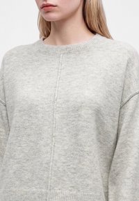 Femme portant un pull tricoté gris clair avec une couture verticale au centre et un col rond, vue des épaules à la poitrine.