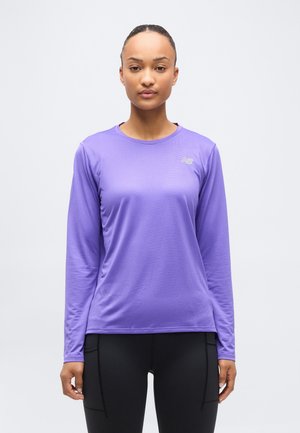 Mujer de pie mirando al frente, vestida con una camiseta deportiva de manga larga morada y leggings negros, sobre un fondo claro y liso.