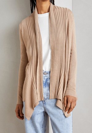 Beige kofta med vertikal ribbning, öppen front och asymmetrisk fåll, i kombination med en vit skjorta och ljus tvättade denimjeans.
