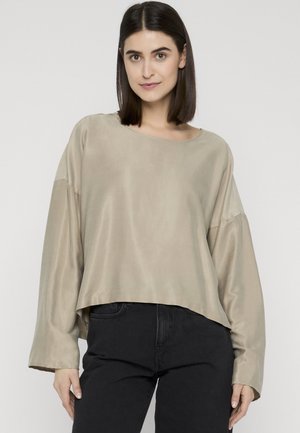 Frau mit glatten dunklen Haaren, die ein lockeres beiges Langarmshirt und schwarze Hosen trägt und vor einem schlichten hellen Hintergrund posiert.