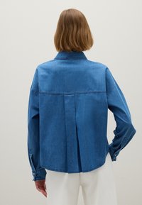 Person mit kurzem braunem Haar, der ein lockeres blaues Jeanhemd und weiße Hosen trägt, von hinten gegen einen schlichten Hintergrund betrachtet.