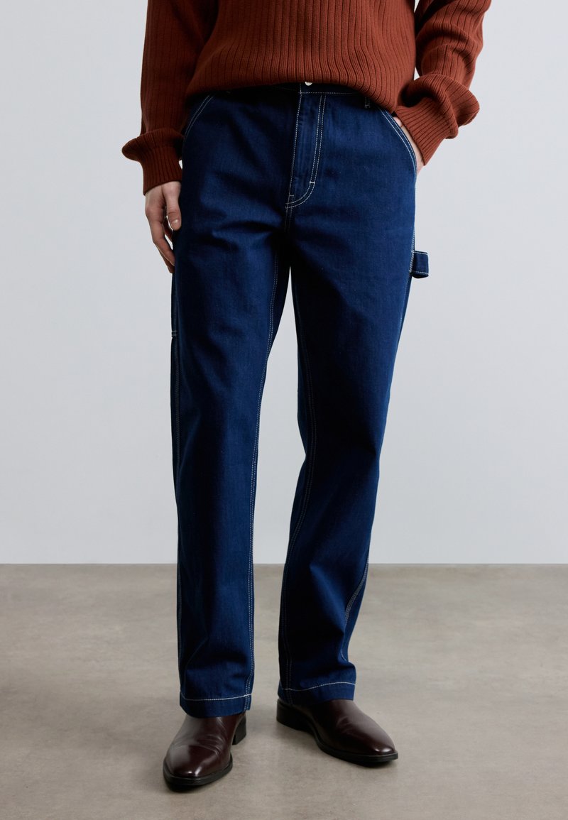 Pantalones vaqueros azul marino con corte recto, costuras en contraste, bolsillos laterales y un lazo en la cintura para accesorios o herramientas.