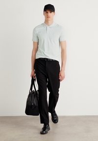 Světle modré polo tričko, černé kalhoty, černé boty, černá kšiltovka. Nese texturovanou černou sportovní tašku, kráčí po neutrální podlaze.