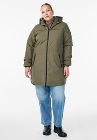 Olīvzaļš pildīts parka mētelis ar rāvējslēdzēju priekšā, kapuci, elastīgām aprocēm un divām sānu kabatām, kombinēts ar gaiši ziliem taisniem džinsiem un melnām zābakiem.