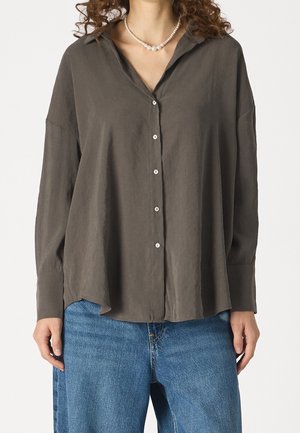 Woman wearing a loose dark gray buttoned shirt with a pearl necklace and blue denim jeans against a plain background.