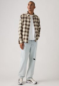 Καρό πουκάμισο με κουμπιά σε μπεζ και καφέ χρώμα, συνδυασμένο με ανοιχτόχρωμο μπλε τζιν με φθορές και ασημένια αθλητικά παπούτσια με μαύρες λεπτομέρειες.