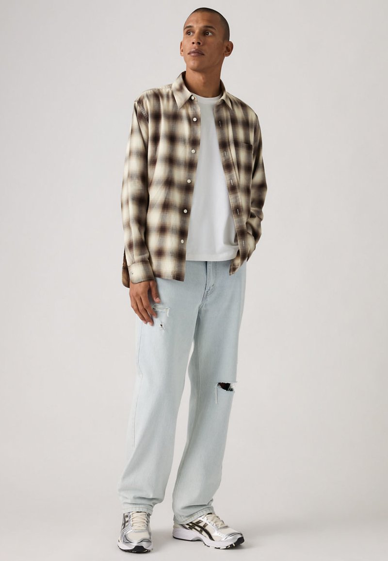 Karierte Hemdjacke in Beige und Braun, kombiniert mit hellblauen, ausgewaschenen Jeans und silbernen Sneakers mit schwarzen Akzenten.