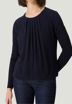 Mujer con una camiseta de manga larga azul marino con pliegues frontales y vaqueros azul oscuro, de pie con una mano detrás de la espalda.