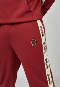 Nahaufnahme einer Person, die rote Hose mit einem weißen Seitenstreifen trägt, auf dem schwarzer "Siksilk"-Text und ein gestickter Wappen-Patch auf dem Oberschenkel zu sehen sind.