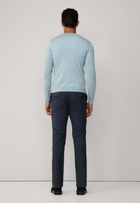 Muž stojí zády, oblečený v světle modrém dlouhém rukávu a tmavě modrých kalhotách s knoflíkovými zadními kapsami na jednobarevném pozadí.