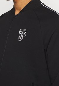 Giacca bomber nera con zip, realizzata in tessuto morbido, con strisce bianche sui lati e un piccolo logo di figura cartoon sul petto.