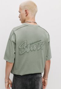 Tricou oversized verde confecționat dintr-un material moale, având un logo texturat și cusut pe spate, cu mâneci scurte și o croială relaxată.