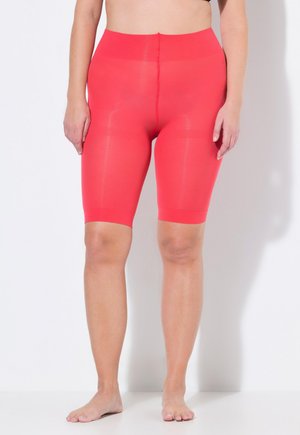 Frau, die leuchtend rote hoch taillierte Kompressionsshorts bis zur Mitte des Oberschenkels trägt, steht barfuß vor einem weißen Hintergrund.