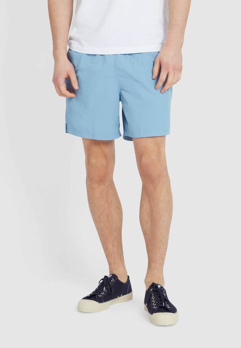 Jasnoniebieskie szorty kąpielowe z elastycznym paskiem w talii, bocznymi kieszeniami i małym akcentem logo. Połączone z ciemnoniebieskimi trampkami z białymi podeszwami.