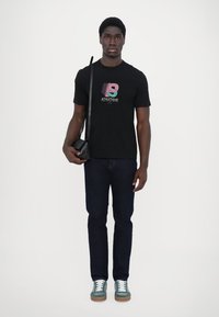Black short-sleeve t-shirt featuring colorful graphic print, paired with dark denim jeans and green sneakers, carrying a small black bag.