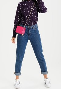 Femme portant un chemisier à pois violets, un jeans bleu retroussé, des baskets blanches et un petit sac bandoulière rose vif.