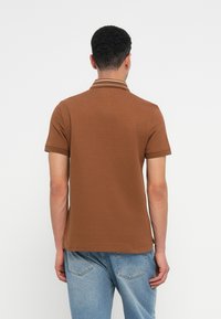 Brūns polo krekls ar teksturētu audumu, īsām piedurknēm un kontrastējošu baltu apkakli. Nēsāts ar gaiši ziliem džinsiem.