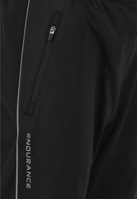 Schwarze Sportleggings mit einer Reißverschlusstasche, ausgestattet mit reflektierenden Akzenten und dem Wort "ENDURANCE" in Weiß entlang der Seite. Weiche Textur.