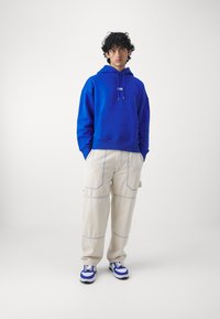 Blå hettegenser med frontlomme, kombinert med beige cargo-bukser med lyse sømmer og blå sneakers med hvite detaljer.