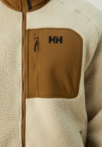 Kurtka polarowa z off-white korpusem, brązową kieszonką na piersi z zamkiem błyskawicznym oraz czarnym logo. Teksturowany materiał z mocnym szwem.
