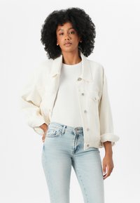 Crème-kleurige spijkerjacket met een ontspannen pasvorm, knoopsluitingen en twee borstzakken, gedragen over een wit shirt en lichtblauwe jeans.