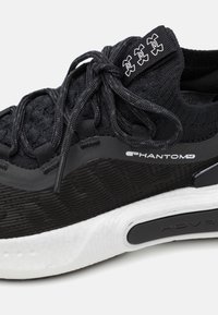 Black athletic shoe featuring a textured knit upper, sleek design, contrasting white sole, and subtle branding elements on the tongue.