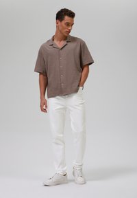 Camicia a maniche corte con bottoni in tessuto marrone testurizzato, abbinata a jeans bianchi a gamba dritta e sneakers bianche, in piedi su uno sfondo neutro.