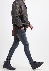 Chaqueta bomber camuflada, vaqueros oscuros y zapatillas negras de caña alta. La chaqueta cuenta con cierre de cremallera y puños acanalados. Vaqueros de corte ajustado con costuras sutiles.