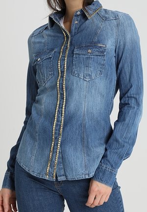 Vrouw draagt een vervaagde blauwe spijkerblouse met borstzakken en decoratieve gouden bies langs de knoopsluiting en kraag, gecombineerd met blauwe spijkerbroek.