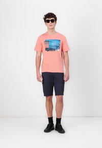 Koralfarvet T-shirt med blåt grafisk design og tekst, parret med marineblå cargo shorts og sorte sneakers, båret med solbriller.