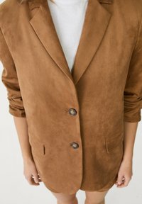 Brauner Wildleder-Blazer mit Reverskragen, zwei Knöpfen und Seitentaschen; besitzt umgeschlagene Ärmel und eine glatte Textur.