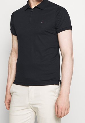 Black polo shirt with a zip neckline, short sleeves, and a small logo detail on the chest, made from a smooth, stretchy fabric.