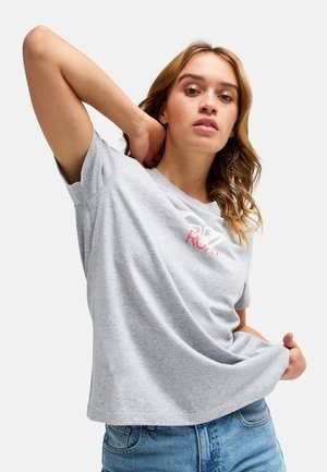 Jeune femme aux cheveux ondulés portant un t-shirt Roxy gris clair et un jean bleu, tenant l'ourlet du t-shirt d'une main, l'autre main derrière la nuque.