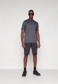 Camisa de homem de mangas curtas com fecho éclair em cinza escuro e detalhes pretos, combinada com calções escuros até ao joelho e sapatilhas pretas. Textura de tecido suave.