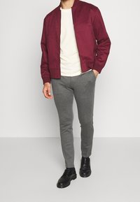 Burgundy bomberjacka, krämfärgad t-shirt, grå avsmalnande byxor och svarta skor. Tyget verkar slätt med minimal detaljering. Åtsittande design.