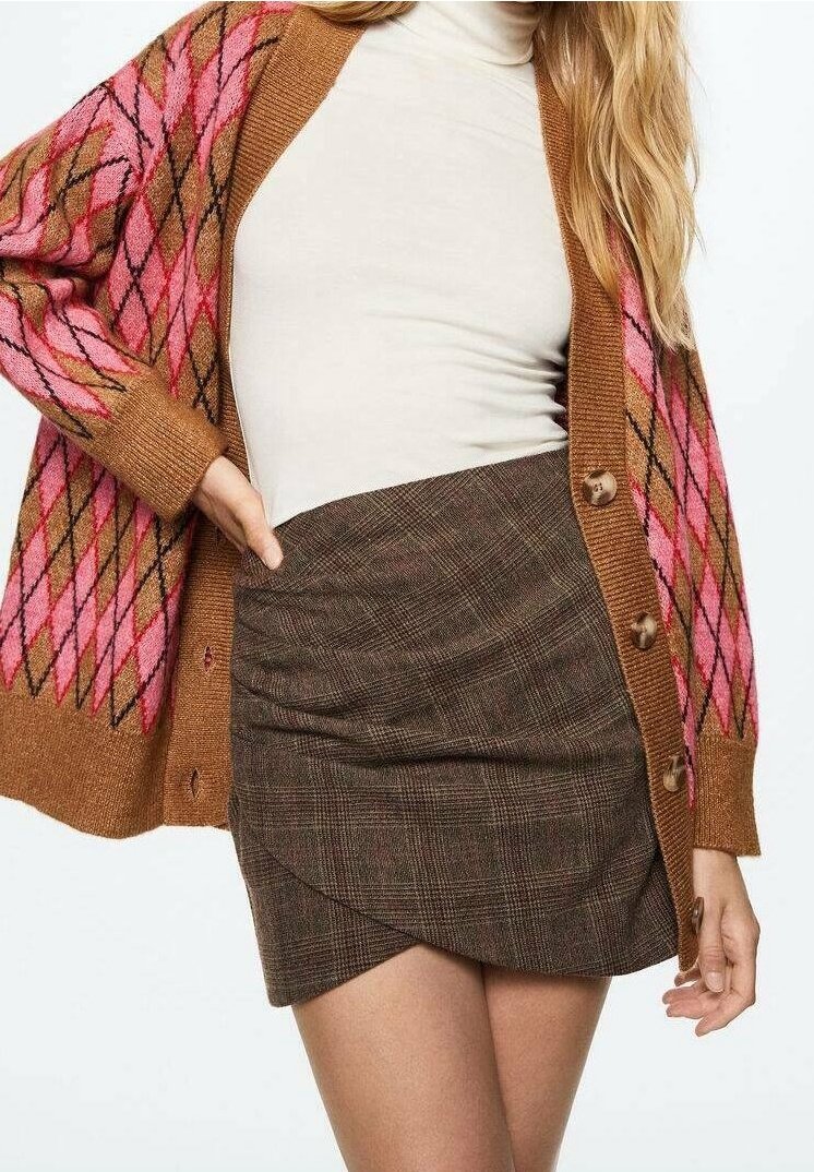 Cardigan en maille marron avec motifs losanges roses et orange, avec boutons. Manches longues, porté sur un col roulé beige et une jupe à carreaux marron.