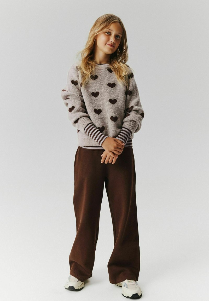 Jeune fille aux longs cheveux blonds debout, portant un pull gris avec des cœurs marron, des poignets rayés, un pantalon marron ample et des baskets blanches.