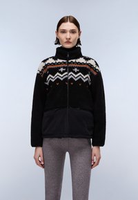 Giacca in pile a fantasia bianca e nera con dettagli arancioni, chiusura con zip, colletto alto e pannello inferiore nero a contrasto. Indossata con leggings grigi.