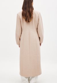 Dlhý béžový kabát z vlnenej zmesi s vypuklými rukávmi, rovnom strihom a švovými detailmi. Tkanina má hladkú, jemnú textúru.