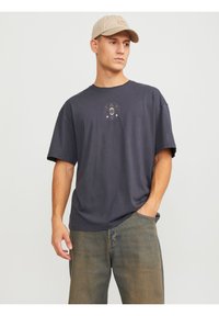 Dunkelgraues T-Shirt mit kurzen Ärmeln und einem kreisförmigen Grafikdesign auf der Brust. Kombiniert mit hellblauen Jeans und einer beigen Mütze.
